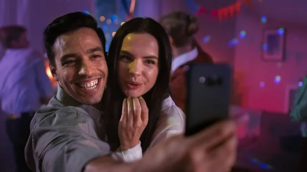 À la Wild House Party : un beau jeune couple prend des selfies avec un smartphone pour le partage de réseaux sociaux. En arrière-plan Une foule de jeunes qui dansent et s'amusent . — Photo