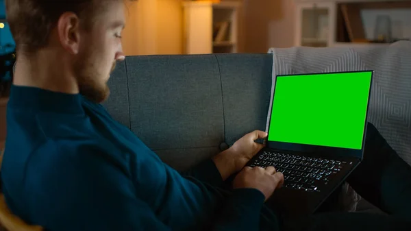 Man Sitting Relaxes on a Couch Works on a Laptop with Green Chroma Key Screen. Late at Night in His Living Room Man Uses Notebook Computer. — Stock Photo, Image