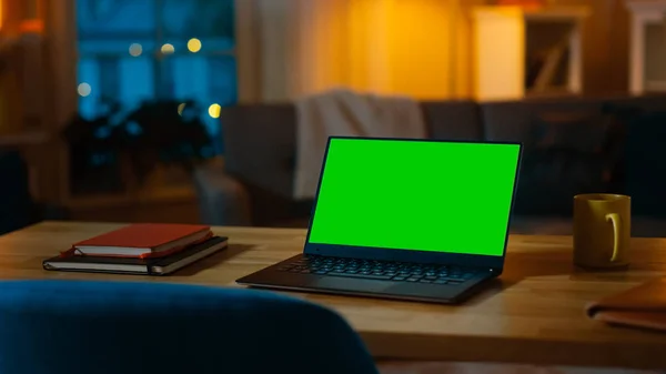Ordenador portátil que muestra la pantalla clave de croma verde se encuentra en un escritorio en la sala de estar. En el fondo acogedor salón por la noche con luces cálidas encendidas . —  Fotos de Stock