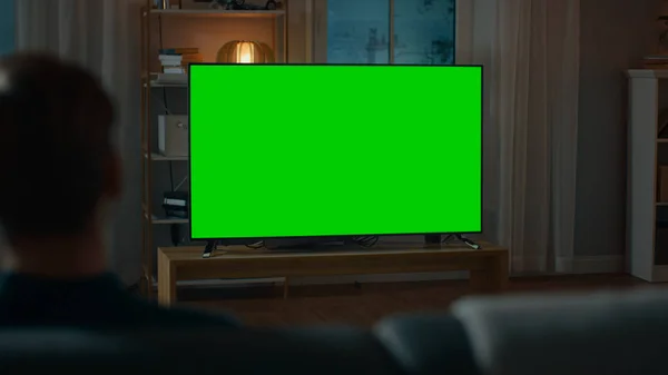 Gece evde bir kanepede otururken adam saatler yeşil mock-up ekran TV. Sıcak ışıklar ile rahat oturma odası. Omuz shot Over. — Stok fotoğraf
