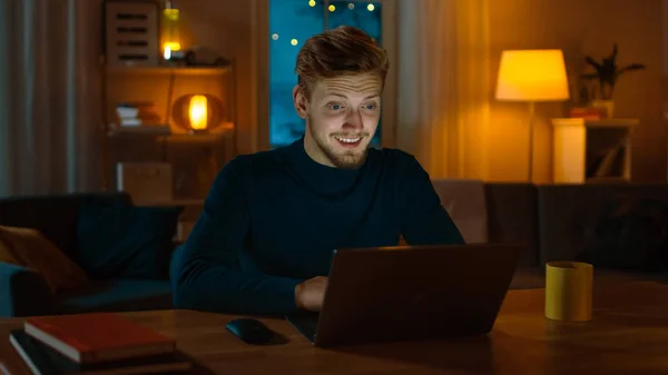 Улыбающийся красавчик работает на ноутбуке, сидя за своим столом дома. Молодой фрилансер работает за компьютером в своей уютной гостиной с горячим вечерним освещением . — стоковое фото