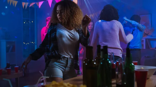The Wild House Party: Beautiful Black Girl Seductively Dances in Neon Lights (engelsk). I bakgrunnen har andre mennesker det gøy og klynger seg . – stockfoto