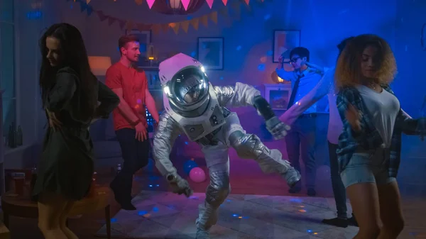 En la fiesta de disfraces de la casa universitaria: el tipo divertido que usa traje espacial baila, hace movimientos modernos de danza robótica. Con él hermosas niñas y niños bailando en luces de neón . —  Fotos de Stock