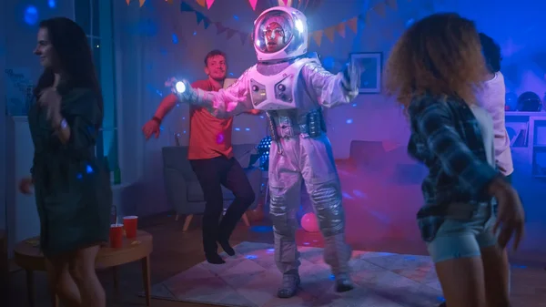 カレッジハウスの衣装パーティーで:宇宙服を着た楽しい男はオフに踊り、ロボットダンスモダンな動きを行います。彼と一緒にネオンライトで踊る美しい女の子と男の子. — ストック写真