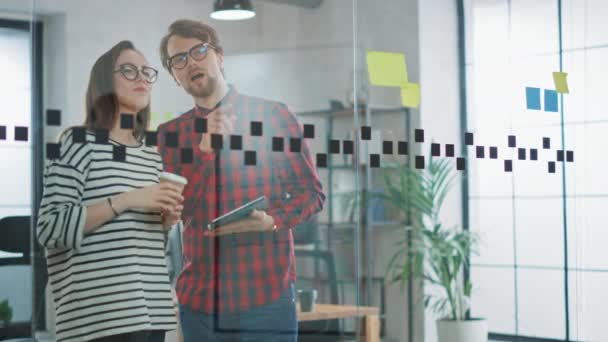 Jeune homme avec tablette et femme avec stand de café à emporter devant un mur de verre avec des notes et discuter des opportunités d'affaires. Ils travaillent à l'Agence Créative. Relations amicales entre collègues . — Video