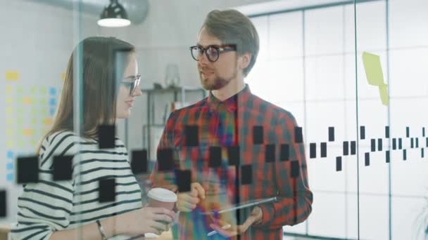 Jonge man met Tablet en vrouw met Afhaalkoffie staan voor een glazen wand met notities en bespreken zakelijke kansen. Ze werken in creatief bureau. High Five over een goed project idee. — Stockvideo