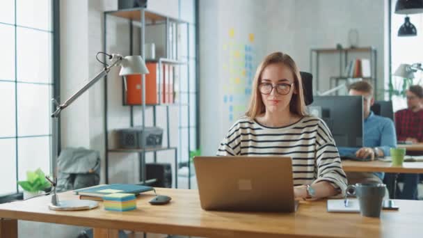 Vacker ung kvinna i glasögon arbetar på en bärbar dator i en kreativ företags byrå. De arbetar i Loft Office. Olika personer som arbetar i bakgrunden. Hon är i en bra vänlig stämning. — Stockvideo