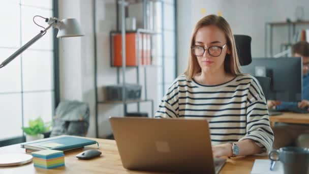 Piękna młoda kobieta w okularach pracuje na laptopie w kreatywnej agencji biznesowej. Pracują w biurze Loft. Zróżnicowana ludzie pracujący w tle. Ona jest w dobrym nastroju friendly. — Wideo stockowe
