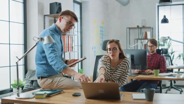 Colega masculina senta-se em uma mesa na frente de uma jovem mulher Cowroker enquanto segurando um computador tablet com notas de negócios. Atmosfera amigável em uma agência criativa. Eles discutem ideias de negócios . — Vídeo de Stock