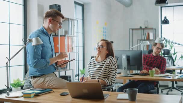 Colega masculina se sienta en una mesa frente a una joven mujer envuelta mientras sostiene una tableta con notas comerciales. Ambiente amistoso en una agencia creativa. Discuten ideas de negocios. — Vídeos de Stock