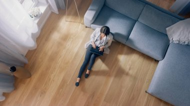 Genç Kadın bir katta oturuyor, laptopta çalışıyor ya da çalışıyor. Modern İçişleri, Gri Kanepe ve Ahşap Döşemeli Konforlu Oturma Odası. Üst Görünüm Kamera Görüntüsü.