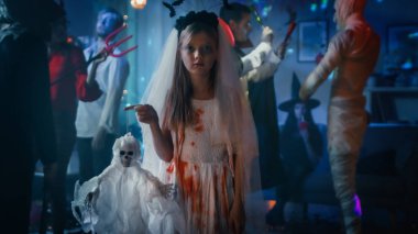 Halloween kostüm parti: Bloody beyaz gelin elbise Holding korkunç bebek küçük kız. Arka plan grubunda canavarlar dans ve disko topu ışık shot ile dekore edilmiş odada eğlenmek