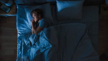 Gece yatak odasında bir yatakta Cozily Sleeping yakışıklı genç adam üst görünümü. Mavi gece renkleri ile soğuk zayıf Lamppost ışık Shining pencere.