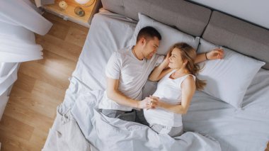 Mutlu genç çift yatağa birlikte sarılarak, Genç kadın hamile ve sevgi dolu ortak dokunur ve onu şefkatli caresses. Üstteki görünüm çekimi.