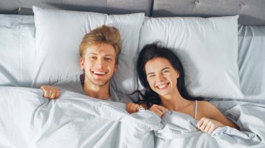 Mutlu ve eğlenceli genç çift battaniye altında bir yatakta, aptal yüzler yapma ve gülüyor yatıyor. Güzel genç Insanlar eğlenmek. Üstten aşağı shot.