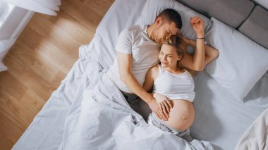 Mutlu genç çift yatağa birlikte sarılarak, Genç kadın hamile ve sevgi dolu ortak dokunur ve onun Belly şefkatli caresses. Güneş parlıyor. Üstten aşağı shot