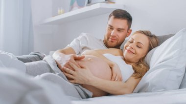Mutlu genç çift yatağa birlikte sarılarak, Genç kadın hamile ve sevgi dolu kocası dokunur ve onun Belly şefkatli caresses. Belly kamera Focus.