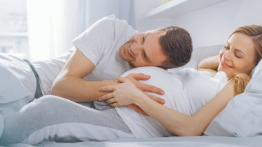 Mutlu genç çift yatakta birlikte cuddling, Genç kadın hamile, sevgi dolu partner eğilir ve bir bebek hareket dinler ve Belly kalp atışı 's. Aile yaşamının ihale ve sevgi dolu anları.