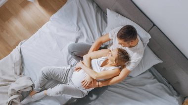 Mutlu genç çift yatakta birlikte sarılmak, Genç kadın hamile ve sevgi dolu kocası kucaklamak ve onun Belly şefkatli okşayan onu tutan
