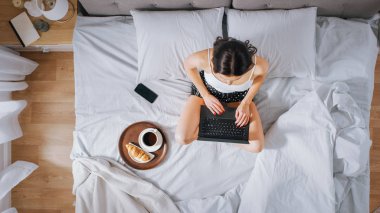 Verimli genç Milenyal kız sabahları bir yatakta oturan, kullanır laptop bilgisayar ve Eats kruvasan ve Içecekler kahve kahvaltı için. Üstten aşağı shot.