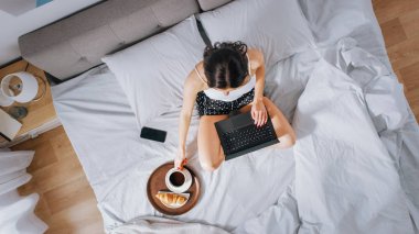 Verimli genç Milenyal kız sabahları bir yatakta oturan, kullanır laptop bilgisayar ve Eats kruvasan ve Içecekler kahve kahvaltı için. Üstten aşağı shot.