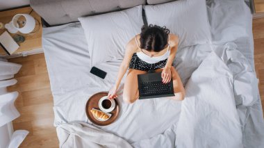 Verimli genç Milenyal kız sabahları bir yatakta oturan, kullanır laptop bilgisayar ve Eats kruvasan ve Içecekler kahve kahvaltı için. Üstten aşağı shot.