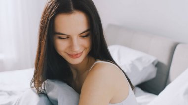 Güzel esmer yavaşça sabah uyanma, yatakta oturur, gülümsemeler charmingly. Güneş büyük pencereden mutlu bir genç kız üzerinde parlar.