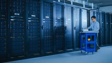 Veri merkezi 'nde: erkek IT teknisyeni çalışan bakım programı bir dizüstü bilgisayar, kontroller operasyonel sunucu raf optimum Işleyişi. Modern High-Tech telekomünikasyon operasyonel süper bilgisayar.