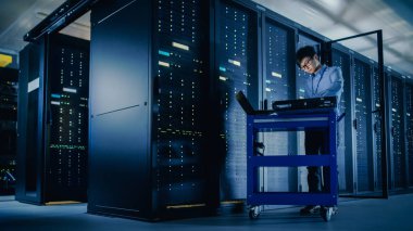Veri merkezi 'nde: erkek IT teknisyeni çalışan bakım programı bir dizüstü bilgisayar, kontroller operasyonel sunucu raf optimum Işleyişi. Modern High-Tech telekomünikasyon operasyonel süper bilgisayar.