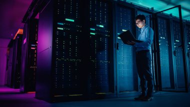 Veri merkezi 'nde: erkek IT teknisyeni çalışan bakım programı bir dizüstü bilgisayar, kontroller operasyonel sunucu raf optimum Işleyişi. Neon renklerde modern High-Tech operasyonel süper bilgisayar, ışıklar