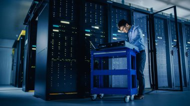 Veri merkezi 'nde: erkek IT teknisyeni çalışan bakım programı bir dizüstü bilgisayar, kontroller operasyonel sunucu raf optimum Işleyişi. Modern High-Tech telekomünikasyon operasyonel süper bilgisayar.