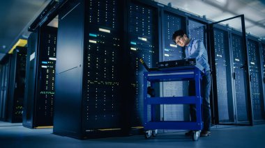 Veri merkezi 'nde: erkek IT teknisyeni çalışan bakım programı bir dizüstü bilgisayar, kontroller operasyonel sunucu raf optimum Işleyişi. Modern High-Tech telekomünikasyon operasyonel süper bilgisayar.