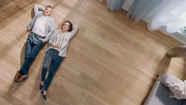 Casal jovem estão deitados em um piso de madeira em um apartamento. Eles são Happy, Smile e Laugh. Sala de estar acolhedora com interior moderno, sofá cinza e parquet de madeira. Top View Camera Shot. — Fotografia de Stock