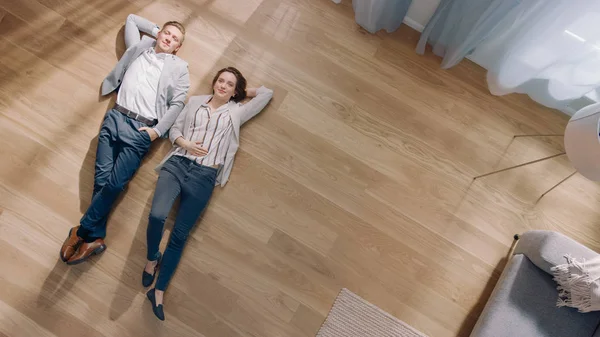 Pareja joven están acostados en un suelo de madera en un apartamento. Son felices, sonríen y ríen. Acogedora sala de estar con interior moderno, sofá gris y parquet de madera. Vista superior de la cámara tiro. — Foto de Stock