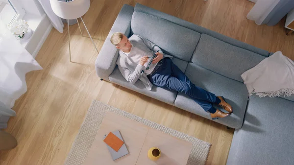 Молодой человек лежит на диване, работает или рисует на планшетном компьютере. Уютная гостиная с современным интерьером, серым диваном и деревянными полами. Фотосъемка сверху. — стоковое фото