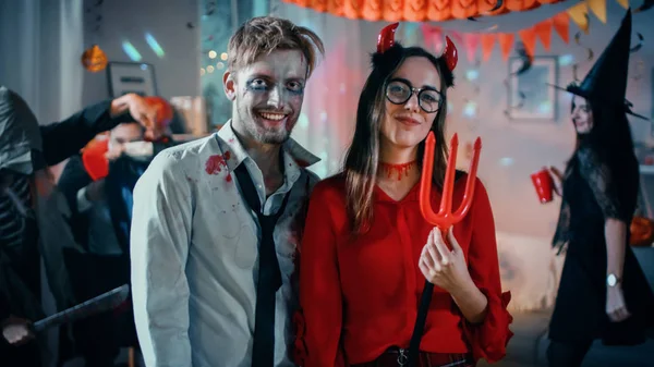 Fête costumée d'Halloween : Zombie mort cérébrale et belle elle diable avec Trident Pose en couple. En arrière-plan Des monstres s'amusent et dansent dans la salle décorée — Photo