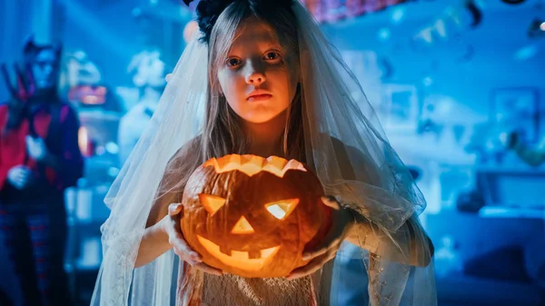 Halloween Costume Party: Portrait of a Little Girl in a Bloody White Bride Dress Holding Burning Pumpkin Head. Zombie, Drácula sedienta de sangre, Momia, Bruja Bruja Bruja y Danza del Diablo Deslumbrante — Foto de Stock