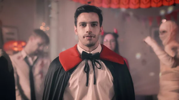 Halloween-Kostümparty: Porträt von Graf Dracula gruselig lächelnd, zeigt seine tödlichen blutigen Zähne. im Hintergrund retro beleuchteter, dekorierter Raum mit schaurigen Monstern, die tanzen — Stockfoto