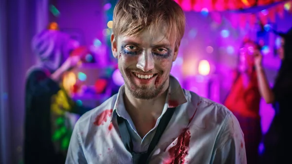 Halloween Costume Party : Portrait of Brain Dead Zombie Wearing Bloody Suit Smiles Creepily. En arrière-plan Des monstres s'amusent et dansent dans la salle décorée — Photo