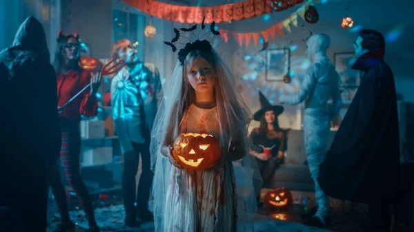 Festa in costume di Halloween: Bambina con un vestito da sposa bianco insanguinato che tiene la bambola spaventosa. Zombie, Dracula assetato di sangue, mamma, strega e abbagliante lei diavolo divertirsi in camera decorata — Foto Stock