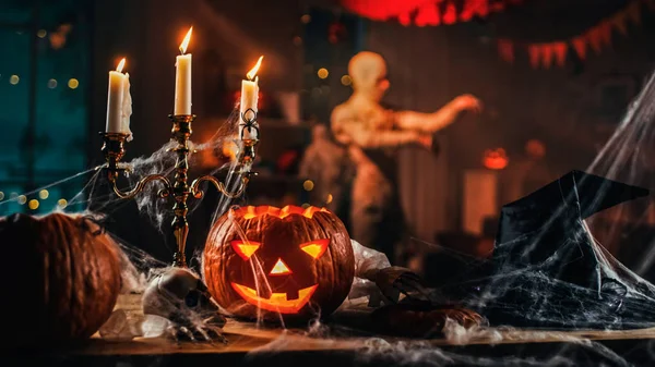 Halloween Bodegón colorido tema: Habitación oscura decorada de miedo con mesa cubierta en telas de araña, calabaza ardiente, Candelabro, Sombrero de bruja y esqueleto. En la silueta de fondo de Monster Walking By — Foto de Stock