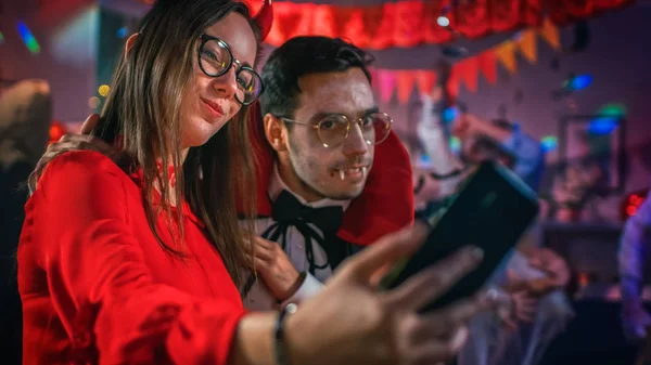 Halloween kostium Party: uwodzicielski diabeł i przystojny hrabia Dracula biorąc selfie dla sieci społecznościowych ze smartfonem. W tle Grupa Monsters zabawy, taniec pod disco ball. — Zdjęcie stockowe