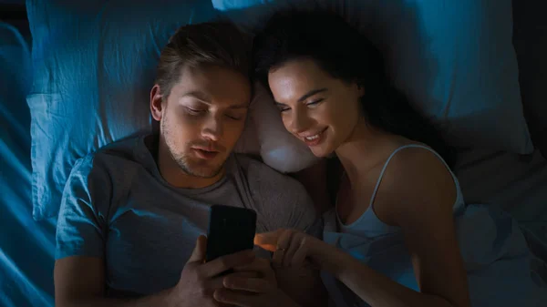 Happy Young Couple Use Smartphone in Bed at Night, They Share Screen, Browsing Through Social Networks, Sharing Pictures, Using Internet and Having Fun. Top View Camera Shot with Blue Colors
