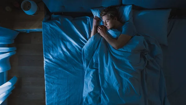Pohled na pohledného mladého muže, který spí Cozily na posteli ve své ložnici v noci. Modré noční barvy se studenou slabou Lamppost zářící skrz okno. — Stock fotografie