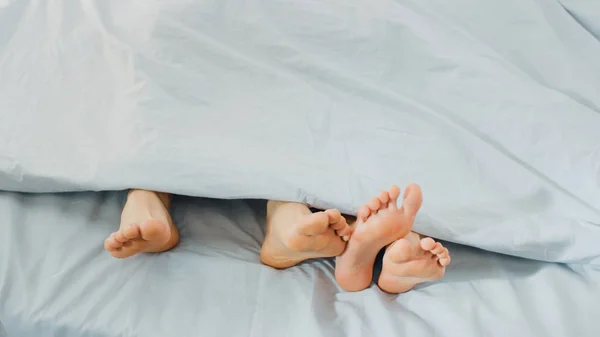 Niedliche Männchen und Weibchen liegen im Bett, die Füße ragen unter der Decke hervor, berühren, flirten, albern herum. helles und gemütliches Schlafzimmer. — Stockfoto