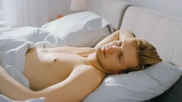 Handsome Fair Boy Lies Cozily in Bed, Slowly Wakes up and Opens His Eyes. Young Caucasian Man. Early Morning Sun Shines Through the Window — Stock Photo, Image