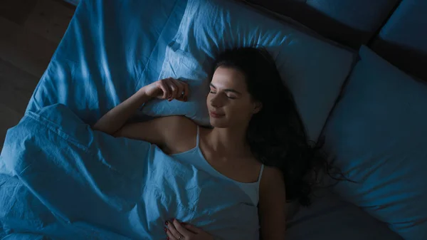 Top pohled na krásnou mladou ženu spící útulně na posteli ve své ložnici v noci. Modrá noční barva se studenou slabostí Lamppost světlo svítí oknem. — Stock fotografie