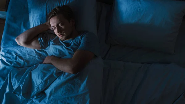 Pohled na pohledného mladého muže, který spí Cozily na posteli ve své ložnici v noci. Modré noční barvy se studenou slabou Lamppost zářící skrz okno. — Stock fotografie