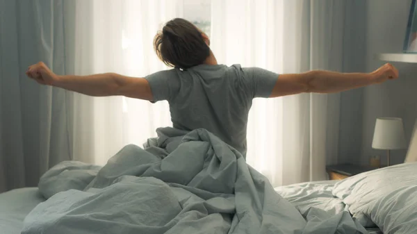 Το όμορφο αγόρι απλώνεται στο κρεβάτι μετά το ξύπνημα. Νέοι και κατάλληλοι Καυκάσιοι άντρες χαιρετά τη νέα μέρα. Ο πρωινός ήλιος λάμπει από το παράθυρο. — Φωτογραφία Αρχείου