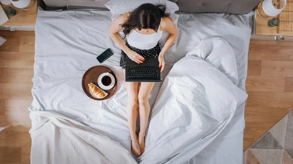Eficiente joven niña milenaria sentada en una cama por la mañana, utiliza ordenador portátil y come cruasanes y bebidas café para el desayuno. Top Down Shot . — Foto de Stock